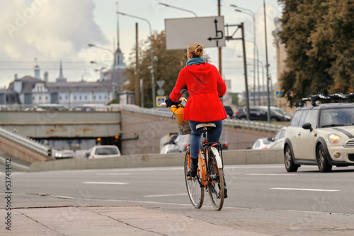 A girl in a red coat rides an orange bicycle around the city next to the auto road.