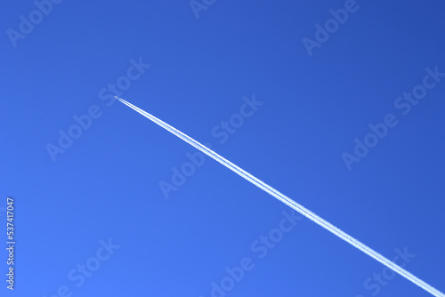 The contrail of the aircraft on the background of a clear blue sky. A distant passenger jet is flying at a high altitude in the blue sky and leaving a long white smoke trail behind it.