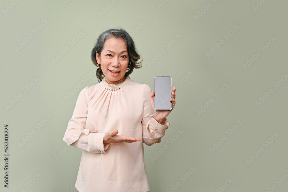 Beautiful 60s senior Asian woman holding a smartphone black screen, green isolated background. Beautiful 60s senior Asian woman holding a smartphone black screen, green isolated background.