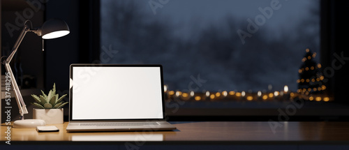 Cozy workspace in Christmas night theme, laptop white screen mockup, table lamp on table