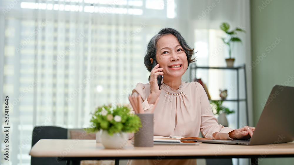 Charming aged-asian businesswoman enjoys talking on the phone while working in her office. Charming aged-asian businesswoman enjoys talking on the phone while working in her office.