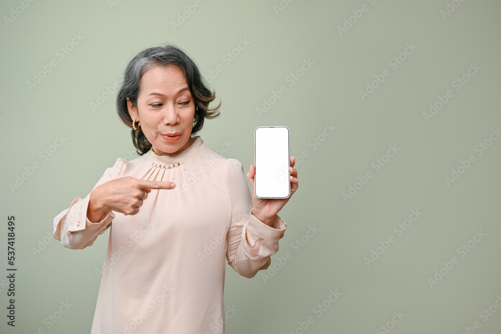 Cheerful aged-asian woman pointing her finger at smartphone, standing against green background