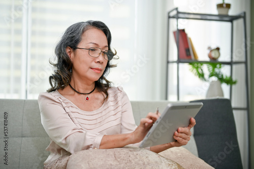 Happy and calm aged-asian woman wearing glasses  using tablet while chilling in living room.
