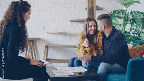 Adorable young couple house buyers are looking at papers and talking with realtor then smiling happily. Beautiful interior and modern furniture are visible.