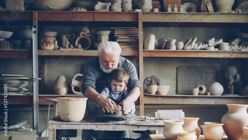 Wallpaper Mural Concentrated small child is forming pot from clay on potter's wheel under guidance of his experienced grandfather. Ceramic pots, vases and figures are in background. Torontodigital.ca