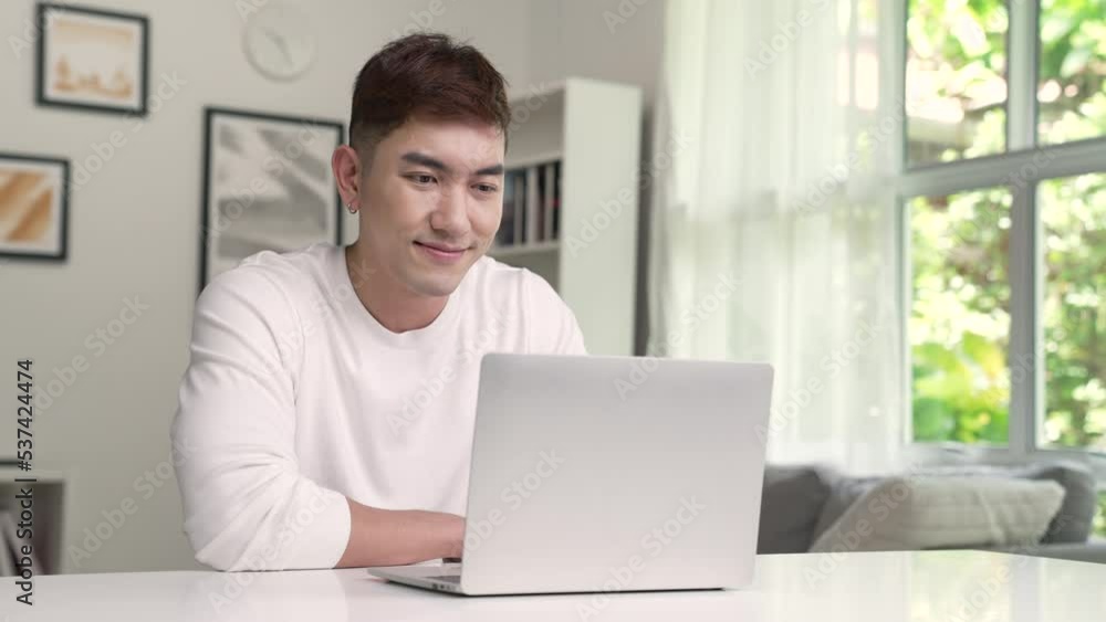 Young Asian man using laptop at home, looking at screen, chatting, reading or writing email, sitting on desk, serious male student doing homework, working on research project online. Young Asian man using laptop at home, looking at screen, chatting, reading or writing email, sitting on desk, serious male student doing homework, working on research project online.