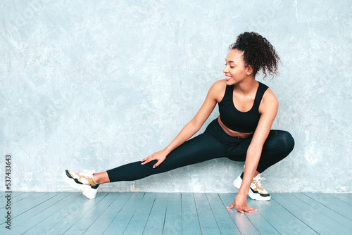 Portrait of fitness smiling black woman in sports clothing with afro curls hairstyle.Wearing sportswear.Sexy young beautiful model doing lunges before training.Female sitting in studio near grey wall