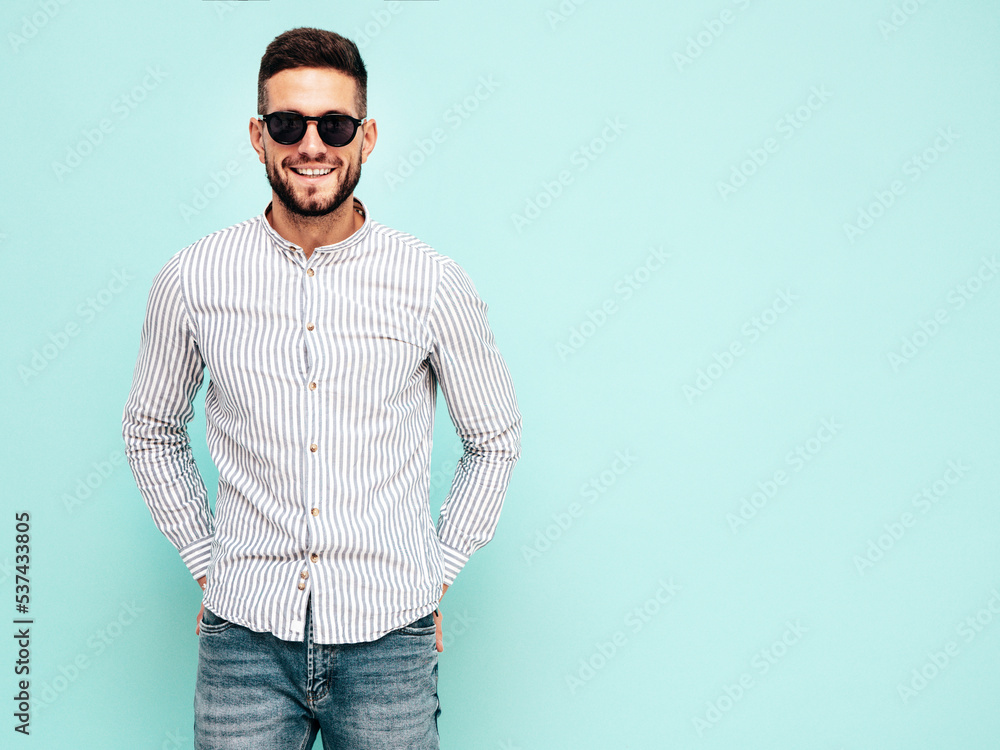 Portrait of handsome smiling model. Sexy stylish man dressed in shirt and jeans. Fashion hipster male posing near blue wall in studio. In sunglasses Portrait of handsome smiling model. Sexy stylish man dressed in shirt and jeans. Fashion hipster male posing near blue wall in studio. In sunglasses