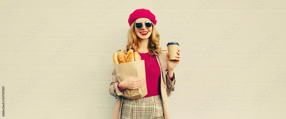 Portrait of happy smiling young woman holding grocery shopping paper bag with long white bread baguette and cup of coffee on gray background, blank copy space for advertising text