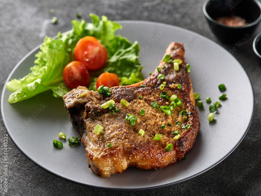 Grilled pork chop on grey plate sprinkled with green onions with salad and cherry tomatoes.