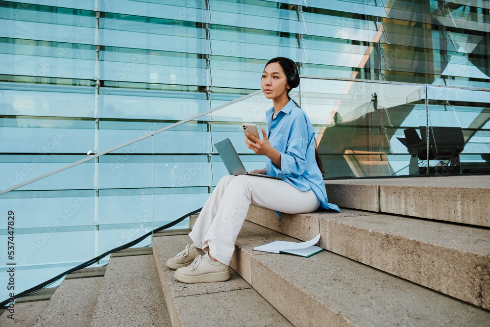 Asian woman in headphones using gadgets while sitting on stairs