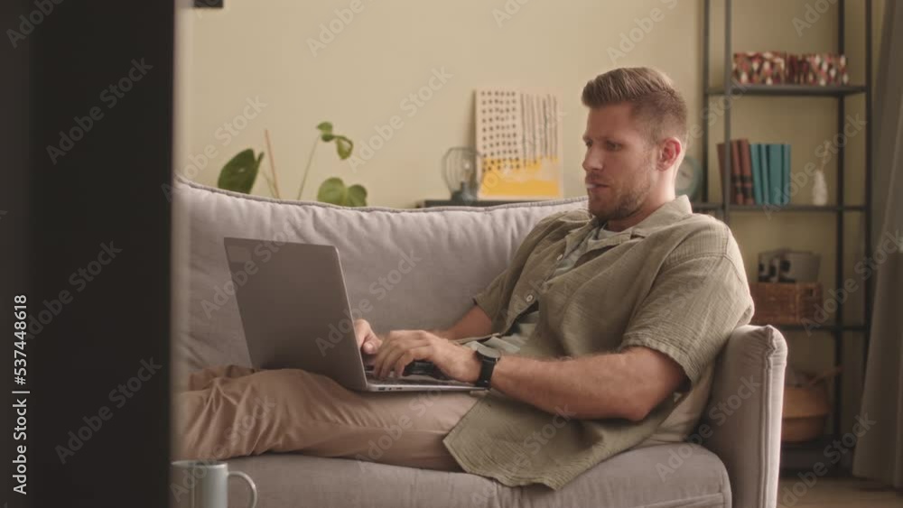 Young Caucasian man sitting relaxedly on sofa in living room vaping and working on laptop Young Caucasian man sitting relaxedly on sofa in living room vaping and working on laptop
