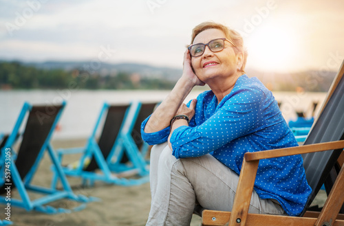 Beautiful happy active senior woman pensioner in a blue shirt sitting resting in a sun lounger on the river bank on the beach