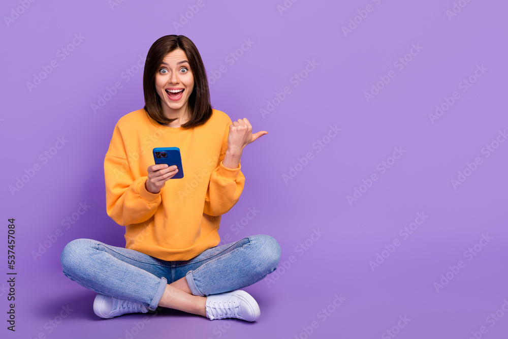 Full body size photo of young woman blogger influencer hold smartphone sitting finger point empty space eshopping promo isolated on violet color background Full body size photo of young woman blogger influencer hold smartphone sitting finger point empty space eshopping promo isolated on violet color background
