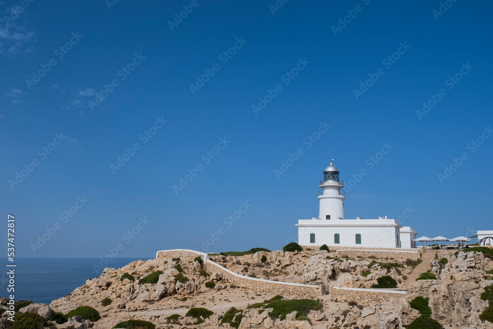 white lighthouse on top of a rocky cliff with white walls surrounding it and the sea in the background, Cavalleria cape lighthouse, Fornells, Menorca Spain white lighthouse on top of a rocky cliff with white walls surrounding it and the sea in the background, Cavalleria cape lighthouse, Fornells, Menorca Spain