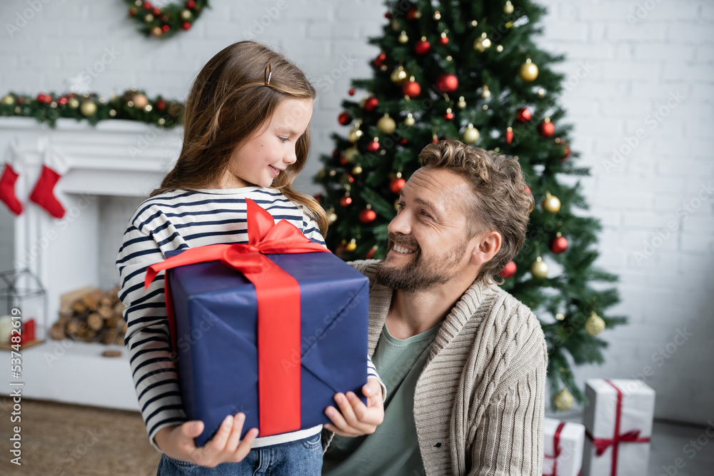 Smiling dad looking at daughter with present near blurred christmas tree at home Smiling dad looking at daughter with present near blurred christmas tree at home