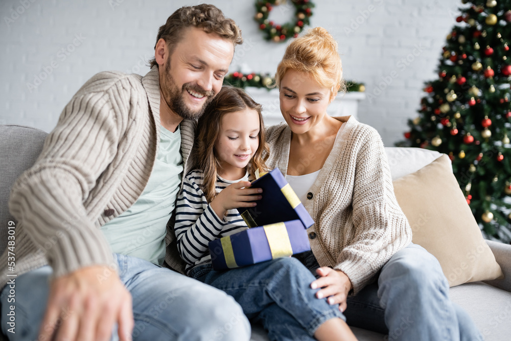 Smiling parents looking at daughter opening christmas gift on couch at home Smiling parents looking at daughter opening christmas gift on couch at home