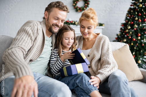 Smiling parents looking at daughter opening christmas gift on couch at home