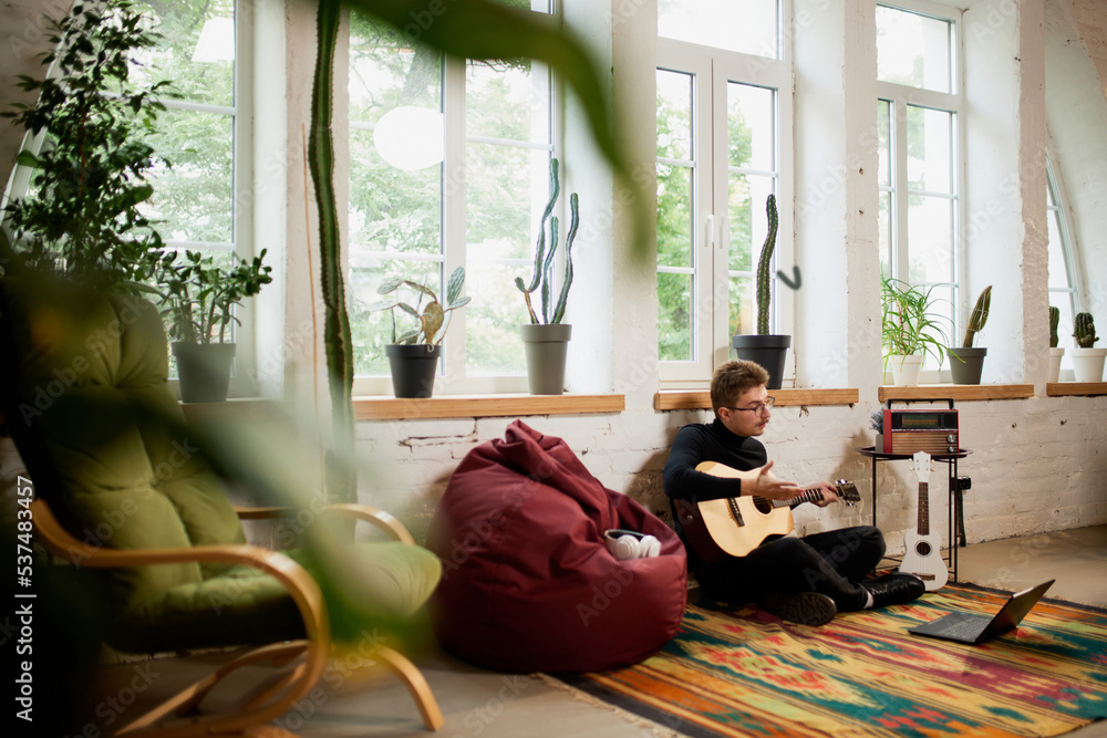 Young man siiting on floor at home and leaning to play guitar. Hobbies and leisure activities. Remote training, online classes