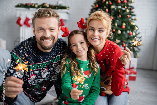 Cheerful family in christmas sweaters holding sparklers and looking at camera at home photo