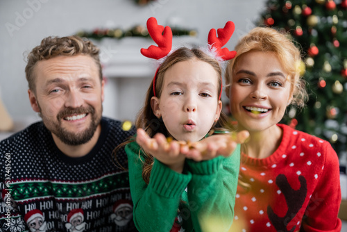 Girl blowing confetti at camera near smiling parents in christmas sweaters at home