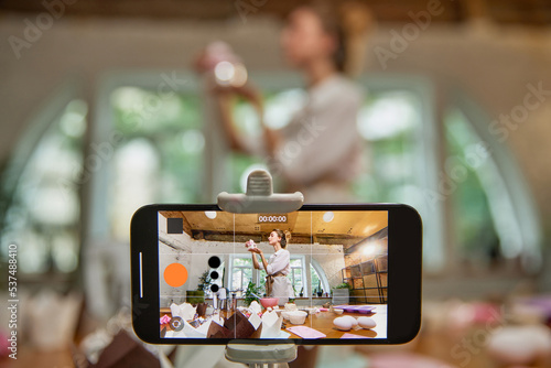 View through phone screen. Young beautiful girl culinary specialist writes video blog on baking. Smiling female vlogger or shooting blog