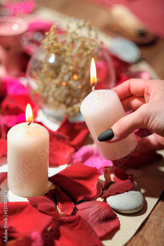  female hand holding burning candle over rose petals, stones and herbs layered on a table. aura readings and magical rituals, spiritualistic and channeling session.