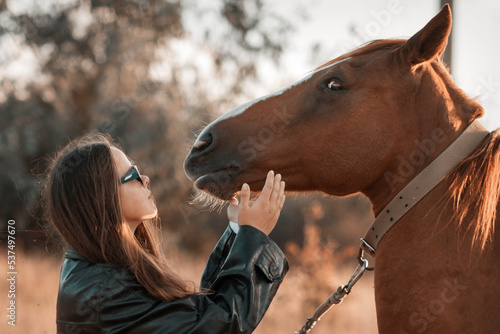 White girl in black leather jacket and glasses next to a horse and poses in the forest at sunset, like in a fairy tale. Well-groomed face with beautiful cosmetics. Enjoy nature and animals. Horse
