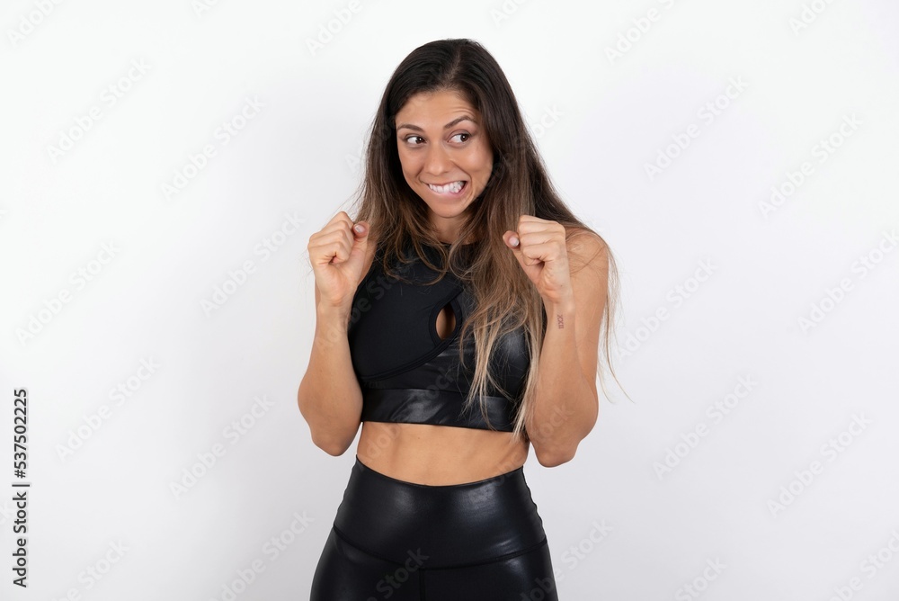 young beautiful woman wearing sportswear over white background clenches fists and awaits for something nice happened looks away bites lips and waits announcement of results young beautiful woman wearing sportswear over white background clenches fists and awaits for something nice happened looks away bites lips and waits announcement of results
