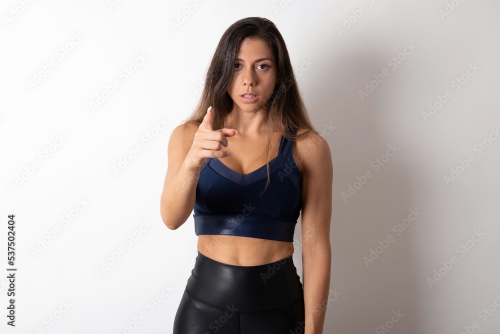 Shocked young beautiful woman wearing sportswear over white background points at you with stunned expression Shocked young beautiful woman wearing sportswear over white background points at you with stunned expression