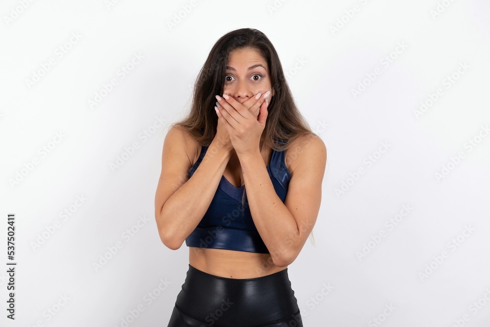 Stunned young beautiful woman wearing sportswear over white background covers both hands on mouth, afraids of something astonishing Stunned young beautiful woman wearing sportswear over white background covers both hands on mouth, afraids of something astonishing
