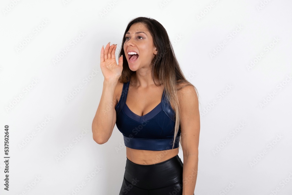 young beautiful woman wearing sportswear over white background look empty space holding hand face and screaming or calling someone. young beautiful woman wearing sportswear over white background look empty space holding hand face and screaming or calling someone.