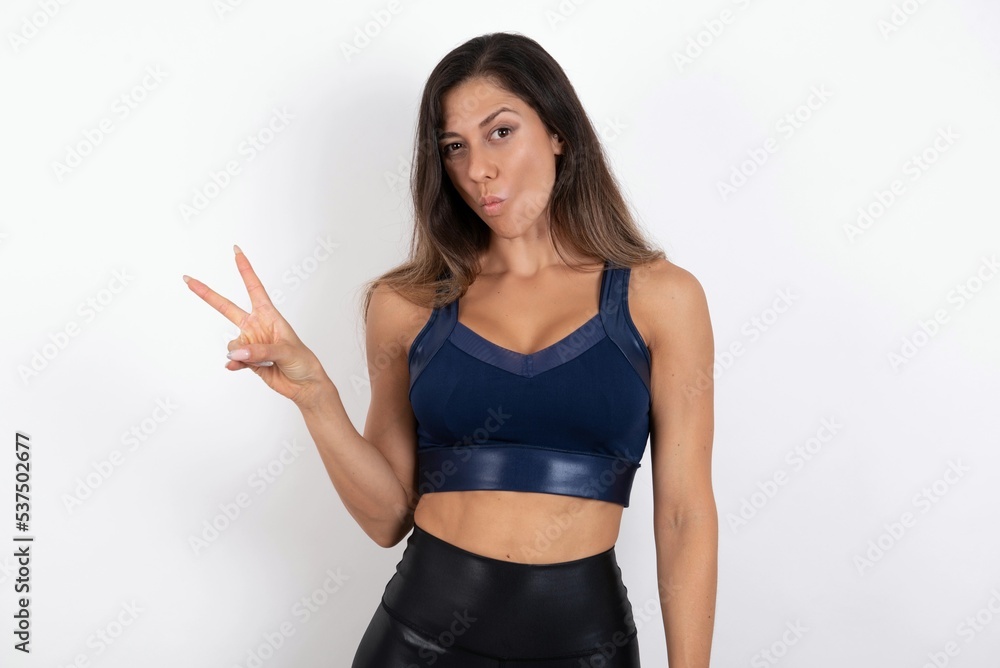 young beautiful woman wearing sportswear over white background makes peace gesture keeps lips folded shows v sign. Body language concept young beautiful woman wearing sportswear over white background makes peace gesture keeps lips folded shows v sign. Body language concept