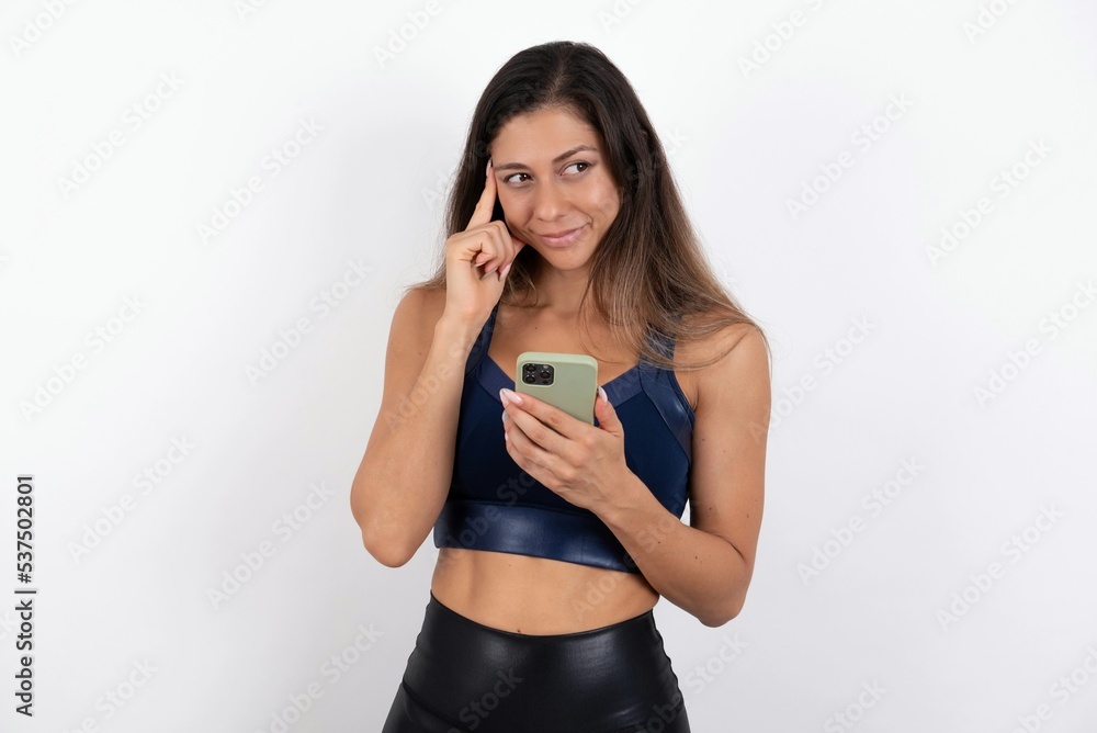 Pensive young beautiful woman wearing sportswear over white background keeps index finger on temple, holds modern smartphone and concentrated aside Pensive young beautiful woman wearing sportswear over white background keeps index finger on temple, holds modern smartphone and concentrated aside