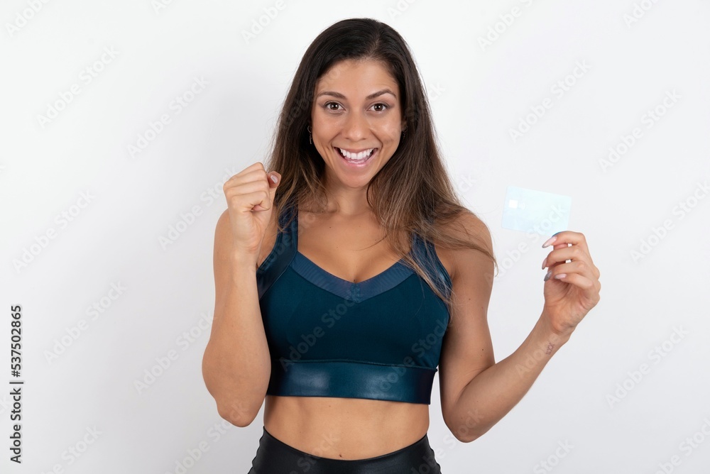 Photo of lucky impressed young beautiful woman wearing sportswear over white background arm fist holding credit card. Celebrated Photo of lucky impressed young beautiful woman wearing sportswear over white background arm fist holding credit card. Celebrated
