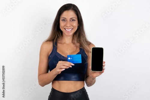 Photo of adorable young beautiful woman wearing sportswear over white background holding credit card and Smartphone. Reserved for online purchases
