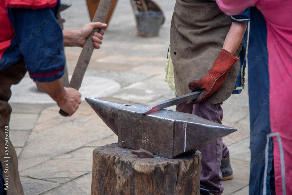 Plano detalle de dos hombres con guantes y delantal elaborando armas y herramientas de manera artesanal sobre un yunque de hierro en el mercado medieval y de artesanía de Hondarribia en el País Vasco. Plano detalle de dos hombres con guantes y delantal elaborando armas y herramientas de manera artesanal sobre un yunque de hierro en el mercado medieval y de artesanía de Hondarribia en el País Vasco.
