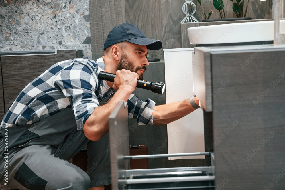 Using flashlight. Plumber in uniform is indoors at home Using flashlight. Plumber in uniform is indoors at home