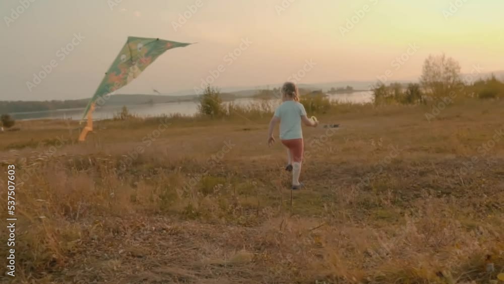 Girl running with a kite in the meadow near the lake. Happy girl playing with kite. Child run with toy on grass in park. Kite soars in wind. Girl with toy kite. Girl playing in park Girl running with a kite in the meadow near the lake. Happy girl playing with kite. Child run with toy on grass in park. Kite soars in wind. Girl with toy kite. Girl playing in park
