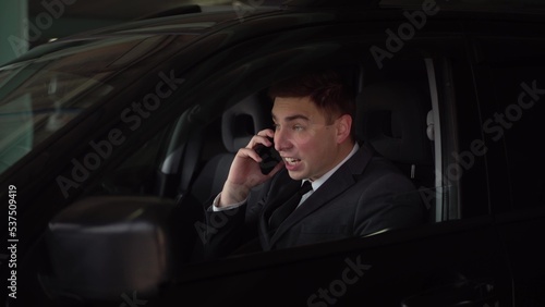 The young boss swears and shouts on the phone while sitting in the car in the parking lot. Angry businessman speaks on the phone. © Vital9c