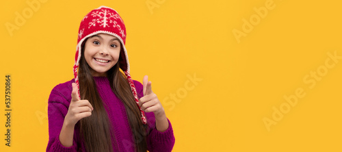 smiling teen girl wear knitted hat pointing finger, keep warm. Child face, horizontal poster, teenager girl isolated portrait, banner with copy space.