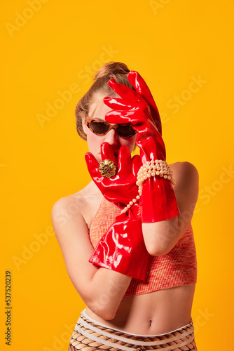 Half-length portrait of young fashionable girl in red huge latex glove and sunglasses sisolated over bright yellow background. Weird people concept. photo