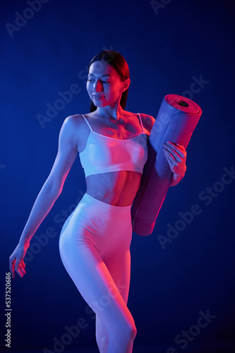 Front view. Holding the mat. Young woman in sportive clothes is in the studio with neon lights