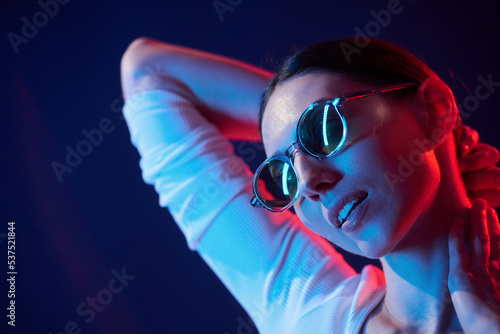 Wearing glasses. Portrait of young woman that is indoors in neon lighting
