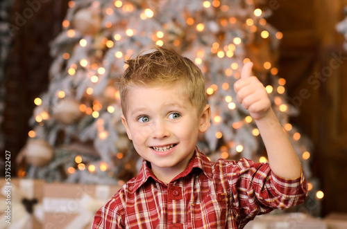 happy blond boy showing thumbs up with his hand, against the background of the New Year's lights of a garland on the Christmas tree