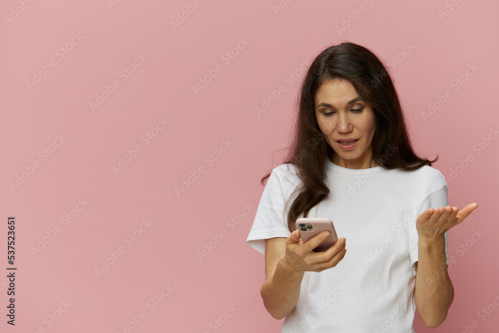 a sad puzzled middle-aged woman stands in a white t-shirt and spreads her hands holding her smartphone. Horizontal photo on a pink background with an empty space for an advertising insert a sad puzzled middle-aged woman stands in a white t-shirt and spreads her hands holding her smartphone. Horizontal photo on a pink background with an empty space for an advertising insert