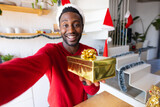 Happy african american man wearing santa claus hat, holding present and having video call