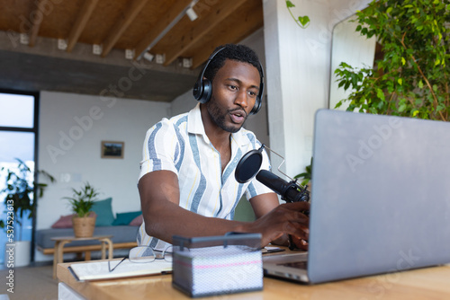 Happy african american man sitting at table in kitchen, using laptop and making vlog photo