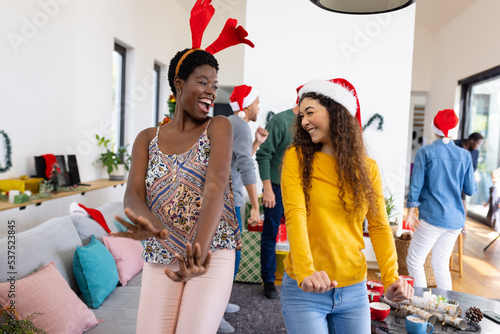 Image of two happy diverse female friends celebrating christmas at home dancing