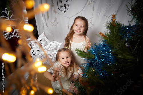 Portrait of a little pretty girls with long beautiful hair in the room decorated for Christmas with garland lights. Christmas and New Year holiday and sisther in white dress as angel photo